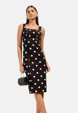Kobieta ubrana w czarną, bezrękawnikową sukienkę midi w białe groszki, trzyma małą czarną torebkę z złotym uchwytem na jednolitym tle.