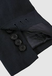 Blazer de culoare bleumarin închis, dintr-un material texturat, având patru nasturi negri și o buzunar ascuns în interiorul clapetei. căptușeală din satin neted vizibilă.