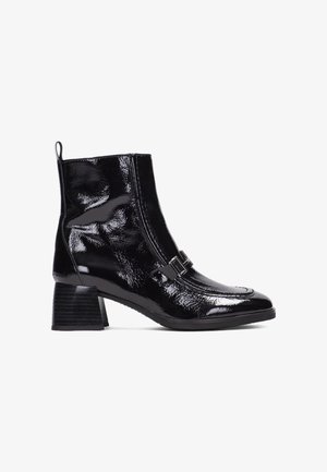 Botín de charol negro con un diseño elegante, puntera cuadrada, cremallera lateral y una hebilla decorativa. Presenta un tacón apilado.