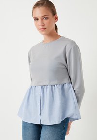 Sivi šik majica dugih rukava s krojenjem na visini struka, koja ima lagani plavi i bijeli prugasti peplum rub i gumbe duž prednje strane.