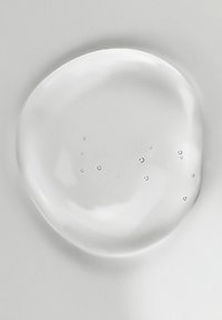 Goccia d'acqua chiara e rotonda su una superficie bianca liscia, con piccole bolle visibili all'interno della goccia, che riflettono leggermente la luce.