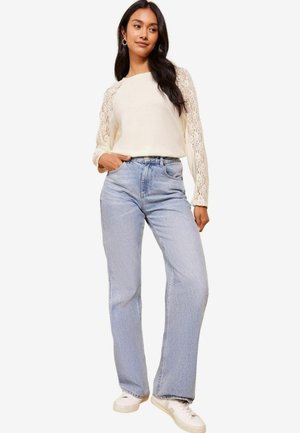 Jeans in denim chiaro con vita alta, gamba ampia e due tasche anteriori, abbinati a una maglia crema con maniche lunghe in pizzo.