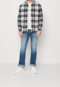 Hombre con una camisa de cuadros en tonos azul marino, blanco y amarillo sobre un suéter blanco jaspeado, jeans azules arremangados y zapatillas blancas, de pie contra un fondo blanco.