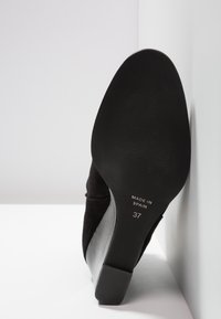 Chaussure à talon haut en daim noir vue de la semelle, présentant une finition texturée, un design effilé et des détails gravés : "FABRIQUÉ EN ESPAGNE" et taille "37".