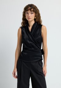 Top sin mangas de satén negro con un diseño de envoltura, que presenta un lazo lateral, textura suave y dobladillo ligeramente curvado. Combinado con pantalones negros.