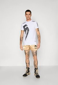 Valge lühikeste varrukatega T-särk musta graafilise logoga, kombineeritud kuldsete satiinist lühikestega, millel on must logo. Mustad saapad beežide detailidega.