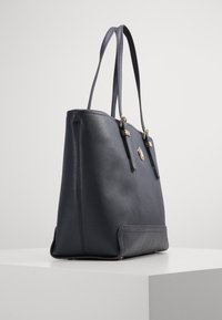 Borsa a spalla in pelle blu navy con doppio manico, hardware dorato e finitura leggermente testurizzata, esposta su un piedistallo bianco.