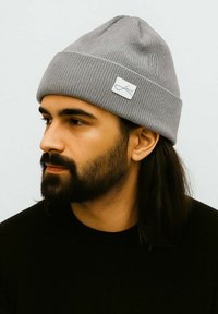 Gray knitted beanie with a ribbed texture, featuring a small white tag with blue lettering. The design is fitted and simple.