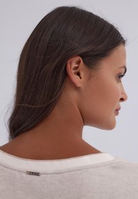 Jasny beżowy sweter o szerokim dekolcie i subtelnej fakturze. Posiada małą metkę z logo z tyłu, prezentowaną na ramieniu modelki.