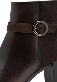 Bottine en cuir marron avec une sangle en daim, boucle métallique circulaire et finition mate. Dotée d'un talon empilé et d'une texture lisse.