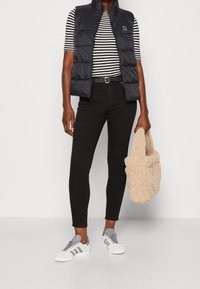 Person, die eine schwarze Steppweste über einem schwarz-weißen gestreiften Shirt, schwarzen Hosen und weißen Sneakers trägt und eine beige, flauschige Tragetasche hält.