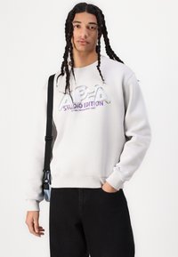 Sweatshirt gris clair avec le texte en relief "ALPHA STUDIO EDITION" en violet et argent. Il a des manches longues et des poignets côtelés ; porté avec un pantalon noir.