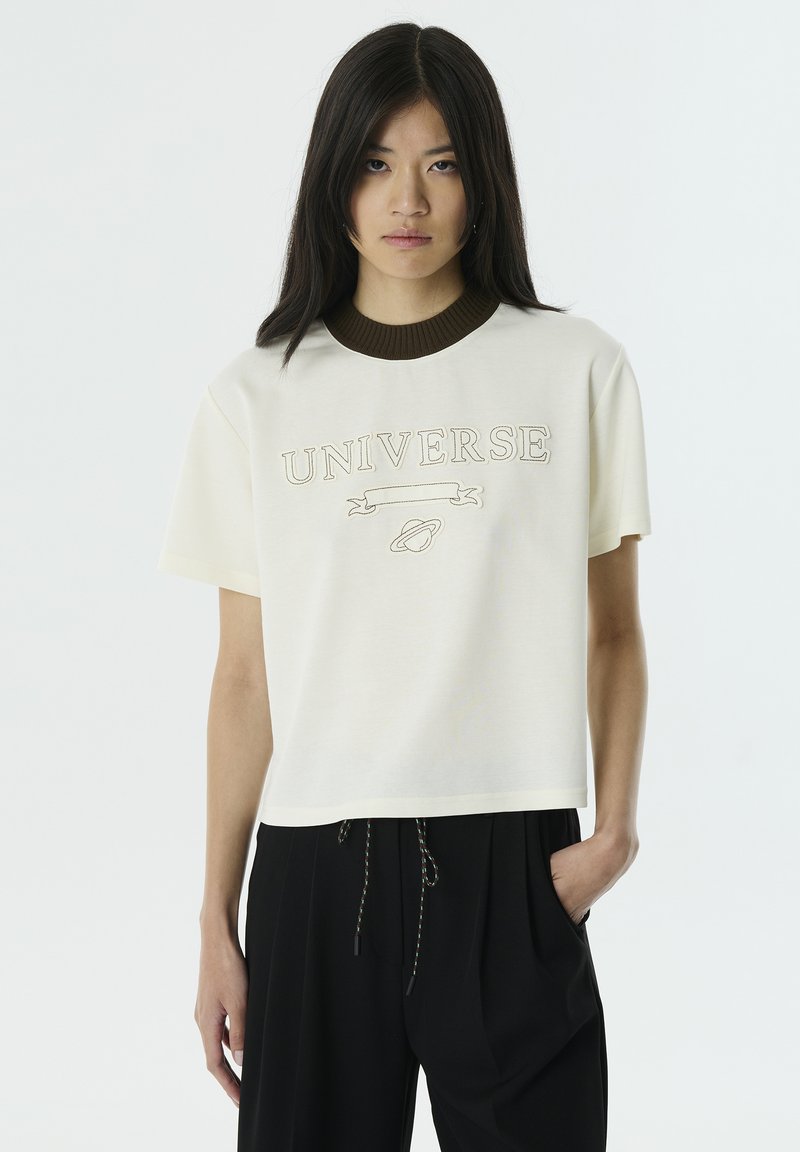 Korte mouwen crème t-shirt met een bruine geribbelde kraag, met geborduurde "UNIVERSE" tekst en een planetenafbeelding op de voorkant. Gladde textuur.