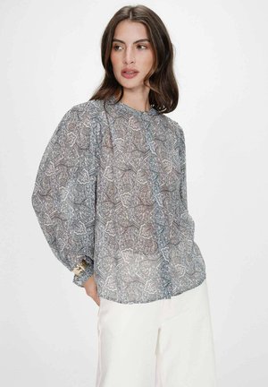 Femme portant un chemisier transparent à manches longues avec motif cachemire et manches bouffantes, associée à un pantalon blanc, debout devant un fond clair uni.