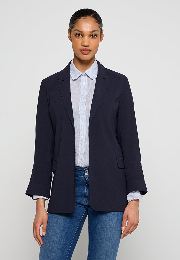 Giovane donna che indossa un blazer blu navy sopra una camicia azzurra a maniche lunghe e jeans blu, in piedi contro uno sfondo chiaro e semplice.