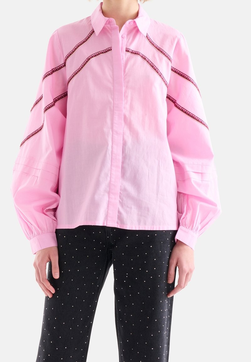 Chemise boutonnée rose clair avec des manches longues bouffantes et des détails brodés rouges contrastants le long des coutures. Associée à un jean noir orné de pois argentés.