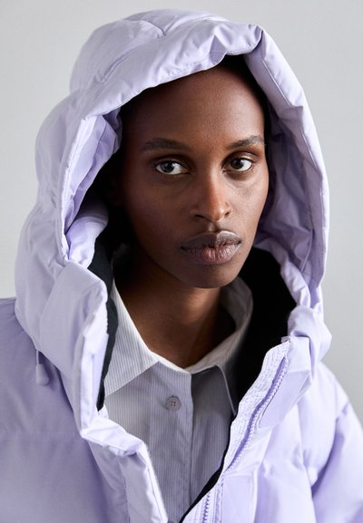 Lilac puffer jacket with a large hood, featuring a black lining and zipper closure, worn over a gray button-up shirt with vertical stripes.