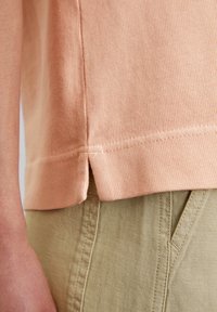 Persikanvärinen ribbineulottu t-paita, jossa on sivuhalkiot helmassa, yhdistetty oliivinvihreisiin housuihin, joissa on teksturoidun pinnan ja näkyvien saumausten yksityiskohdat.