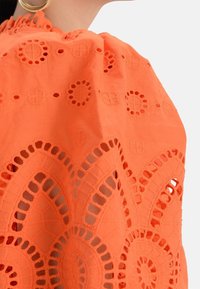 Vrouw in een oranje top van gehaakt borduurwerk met pofmouwen en gouden ringoorbellen, geknipt bij de schouders en nek.