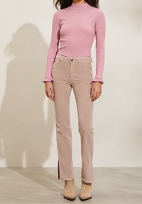 Femme portant un haut à manches longues rose côtelé, un pantalon beige taille haute avec fentes latérales, et des bottines en suede beige, debout sur un sol beige.