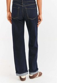 Zadní pohled na osobu oblečenou v tmavě modrých ohrnutých džínách a hnědých botách s bílými kotníkovými ponožkami na bílém pozadí.