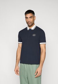 Μπλε ναυτικό πόλο με λευκές λεπτομέρειες και λογότυπο στην αριστερή πλευρά του στήθους· κατασκευασμένο από απαλό ύφασμα με κλασικό γιακά και κοντά μανίκια.