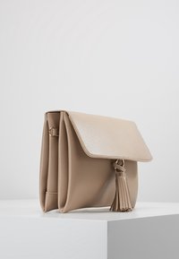 Borsa a mano in pelle beige con superficie texturizzata, chiusura a pattina e un elegante pon-pon decorativo. Presenta un design compatto e quadrato con una leggera lucentezza.