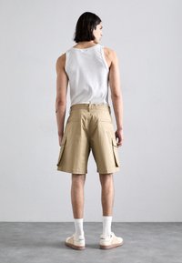 Pantalones cortos cargo color beige con bolsillos laterales, combinados con una camiseta sin mangas blanca y calcetines deportivos blancos. Tejido suave, ajuste relajado. Fondo liso.