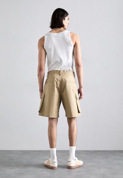 Tan cargo shorts with side pockets, paired with a white tank top and white athletic socks. Smooth fabric, relaxed fit. Plain background.