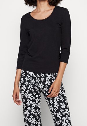 Femme portant un haut noir à manches longues avec un col festonné et un pantalon noir orné d'un motif floral blanc, debout devant un fond uni.