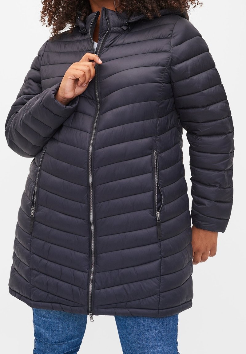 Nafouklý černý kabát s horizontálním prošíváním, zapínání na zip a dvěma bočními kapsami. Má kapuci a je vyroben z lesklého, hladkého materiálu.
