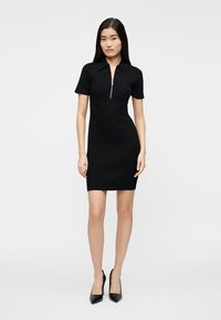 Črna rebrasta obleka s kratkimi rokavi in z zadrgo na izrezu. Gladek tekstura in prilegajoč se kroj, kombinirano s črnimi visokimi petami.