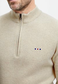 Beige zipzáras pulóver texturált anyagból. Bordás gallérral rendelkezik, és egy kis hímes mintával díszítették kék, piros és fehér színekkel.