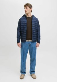Jachetă puffer bleumarin cu glugă, textură matlasată. Purtați-o peste un pulover din lână cu model de sârmă brun, asortată cu blugi drepți de un albastru deschis și încălțăminte bej.