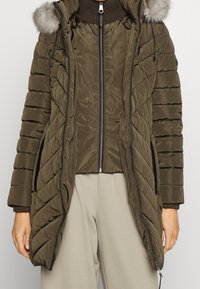 Olivgrüner gesteppter Wintermantel mit Fellkapuze, Reißverschluss, gerippten Bündchen und Seitentaschen, getragen über beigen Hosen.