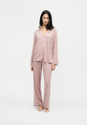 Femme debout pieds nus portant un haut ample à boutons rose clair à manches longues et un pantalon de pyjama large assorti sur fond blanc.