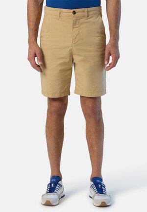 Pantaloni corti beige realizzati in tessuto leggero con parte anteriore piatta, tasche standard e chiusura con bottone. Indossati con scarpe da ginnastica blu.