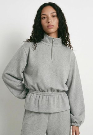 Ung kvinde med krøllet hår iført en lysegrå sweatshirt med halv lynlås og matchende bukser, stående mod en ensfarvet baggrund.