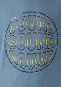 Lichtblauwe katoenen t-shirt met een ronde afbeelding en grote, vetgedrukte tekst "SOUND" in zwart en geel. Inclusief extra tekst in een draaiend patroon.