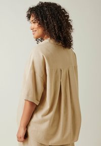 Camicia di lino beige con vestibilità ampia, spalle ribassate e dettaglio di piega sul retro. La texture morbida e le maniche corte aumentano il comfort.