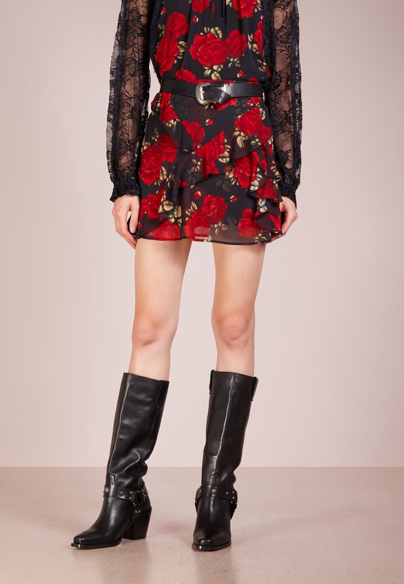 Botas de cuero negras hasta la rodilla con un tacón grueso y correas decorativas, combinadas con un vestido negro de volantes con estampado floral y un cinturón negro.