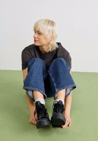 Bottes à plateforme noires avec lacets, jeans larges en denim et t-shirt sombre, assis sur un sol vert clair. Arrière-plan minimal, tons neutres.
