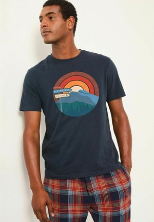 Marineblaues Baumwoll-T-Shirt mit grafischem Runddruck, das einen Sonnenuntergang, Berge und Wegweiser zeigt. Kurze Ärmel, lockere Passform. Kombiniert mit karierter Shorts.