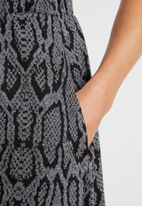 Ruka umístěná uvnitř kapsy černého a šedého vzorovaného oděvu s abstraktním geometrickým designem, detailní pohled.