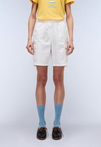 Weiße Cargo-Shorts mit Seitentaschen, kombiniert mit blauen Knöchelsocken und braunen Schuhen. Das Outfit umfasst ein gelbes T-Shirt mit einem Grafikdesign.