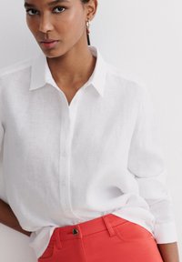 Camicia bianca in lino con colletto, abbinata a pantaloni rossi a vita alta con dettaglio di bottone in metallo. Tessuto testurizzato visibile.
