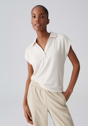 Junge Frau trägt ein ärmelloses weißes Oberteil mit Kragen und beige Hose, posiert mit einer Hand in der Tasche vor einem schlichten hellen Hintergrund.