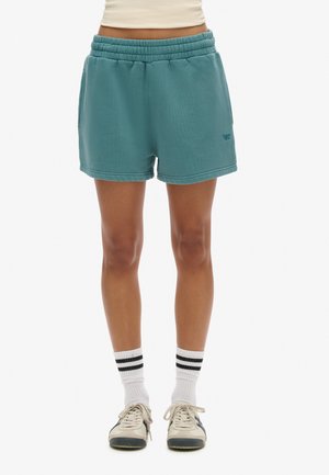 Personne portant un short à taille élastique bleu sarcelle, des chaussettes blanches avec des rayures noires et des baskets blanches, debout devant un fond blanc.