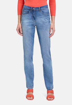 Kvinde iført lyseblå jeans med lige ben, orange langærmet top og orange flettet rem åbne tå hæle, står mod hvid baggrund.