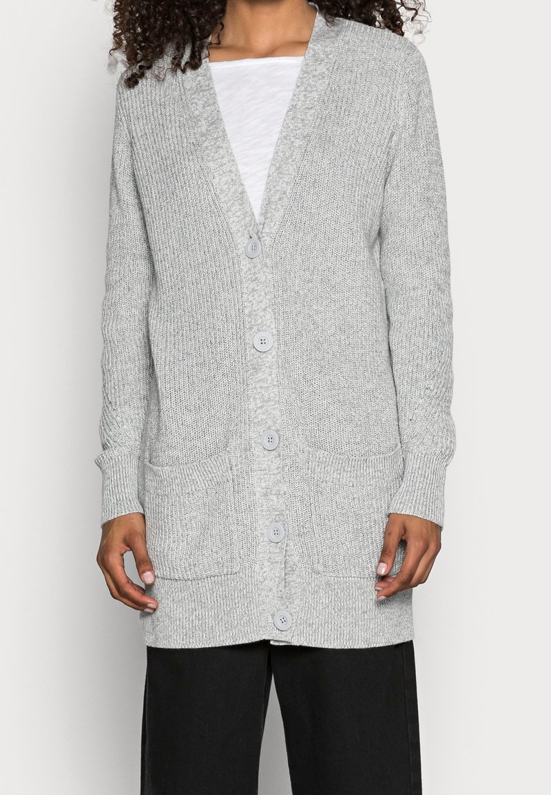 Světle šedý pletený cardigan s výstřihem do V, šesti knoflíky, dvěma předními kapsami a žebrovanými manžetami a lemem. Měkká textura a uvolněný střih.